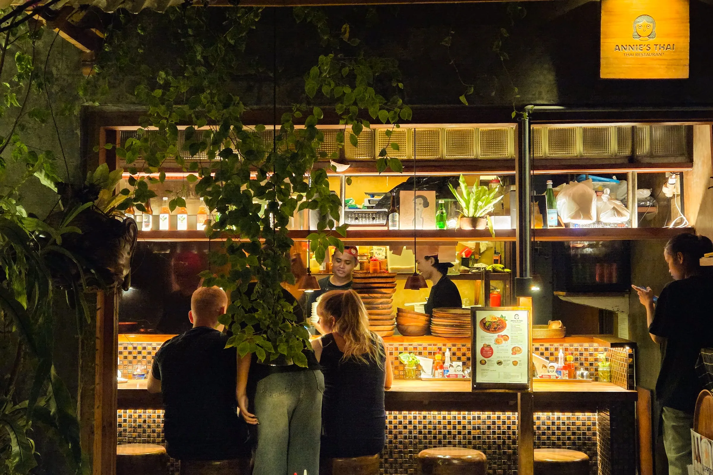 people standing in front of a bar at night with lights on and plants growing out of the shelves