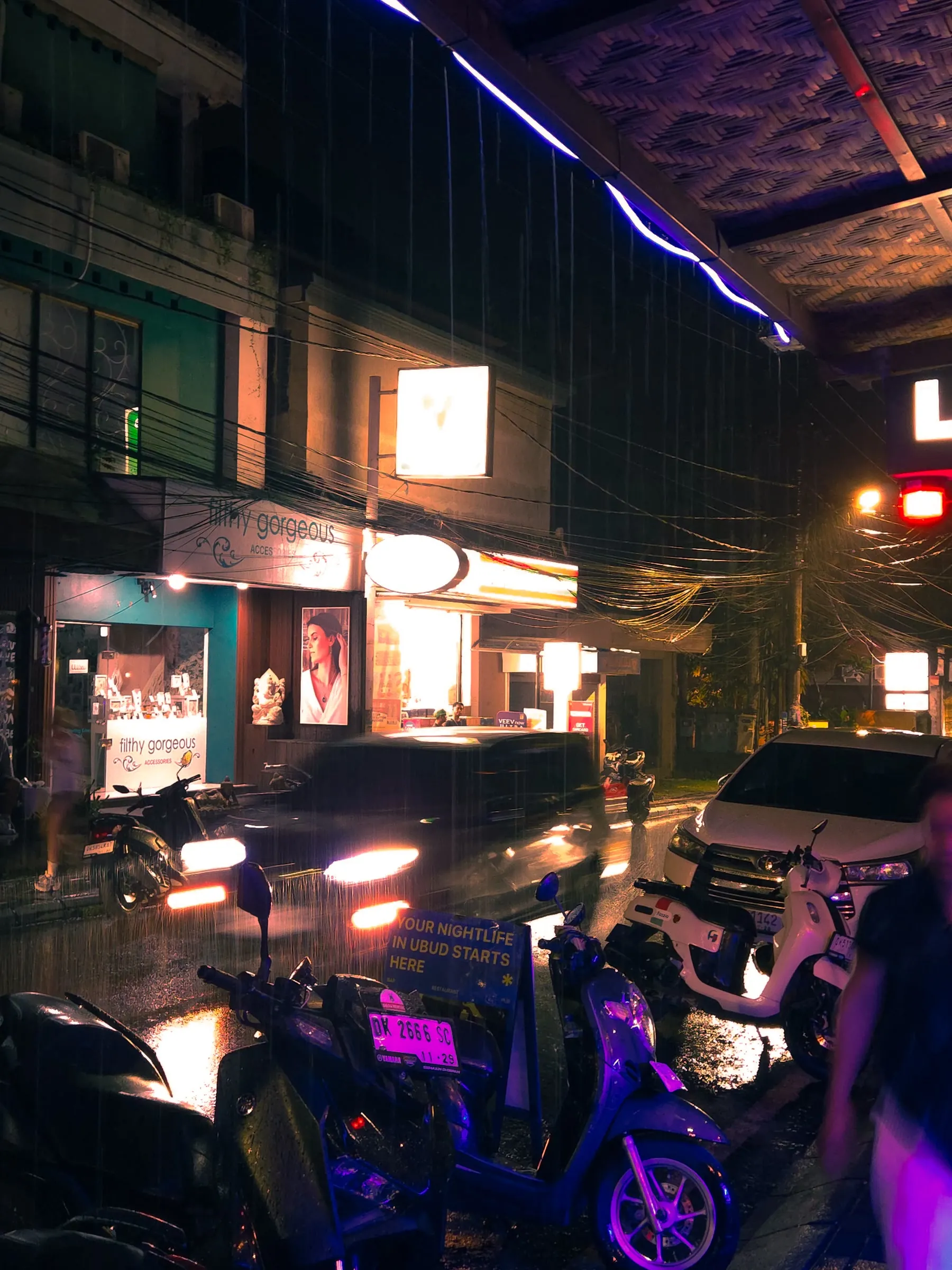 a city street at night with parked motorcycles and people walking on the sidewalk in the rain